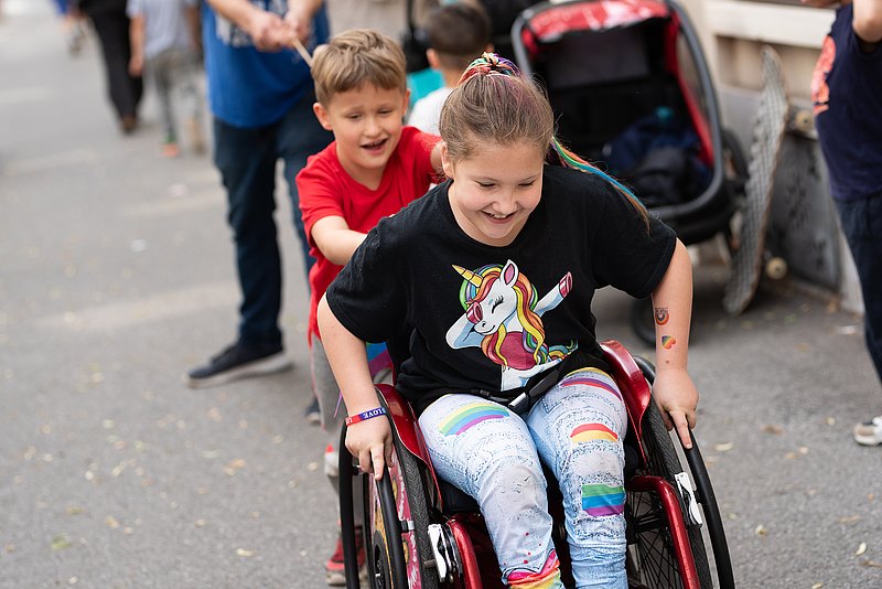 Kinderaktiv - Hollis Grätzltour Ein Kind im Rollstuhl, lachend, mit den Händen an den Rädern. Ein Kind läuft dahinter und hat die Hände am Rollstuhl
