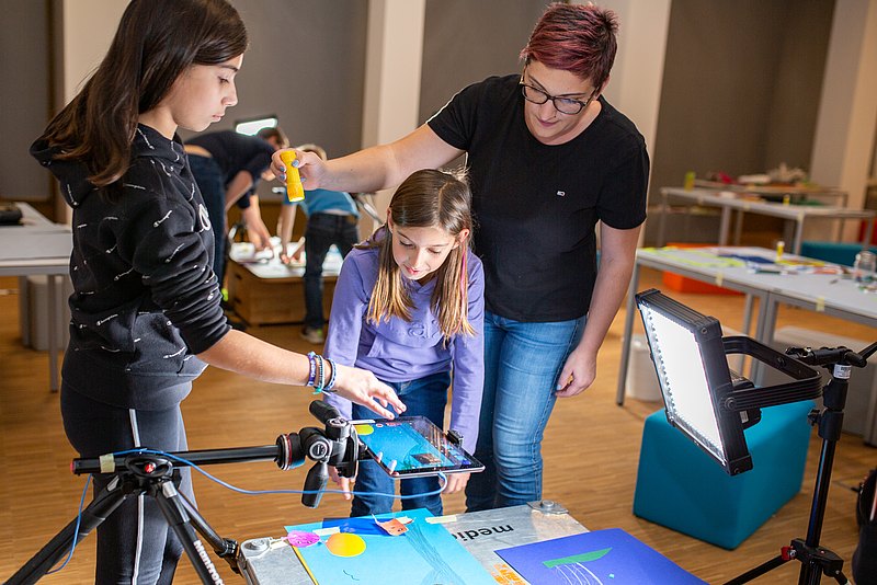 _Q4A5658_XLargeJPG Zwei Kinder/Jugendliche schauen von oben auf einen Screen. Eine erwachsene Person steht daneben und leuchtet mit einer Taschenlampe von oben auf den Screen