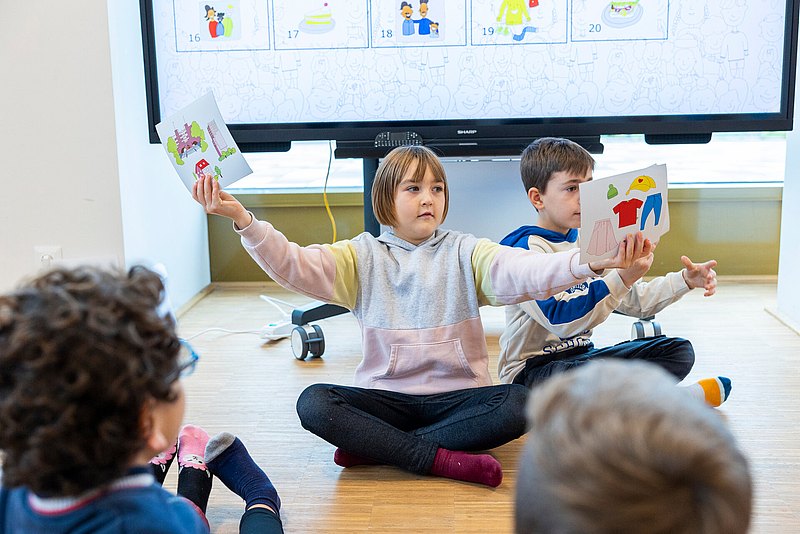 221117_0891 Am Boden sitzen Kinder, eins davon hat Bildtafeln in der Hand und hät sie mit weit ausgestreckten Händen