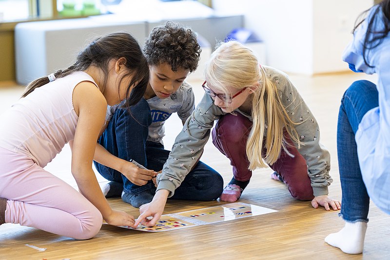 221117_1735 Drei Kinder sitzen am Boden vor einem Spiel und schauen und zeigen auf die Unterlage